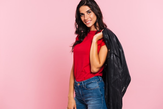 Vista laterale del rivestimento sorridente della tenuta della donna del brunette sulla spalla e sull'esame della macchina fotografica sopra il colore rosa