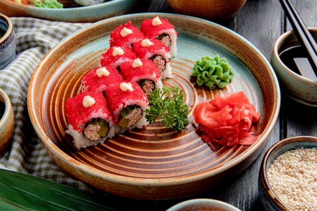 Vista laterale del rotolo di sushi con l'avocado del granchio coperto di caviale rosso con zenzero e wasabi su un piatto su legno