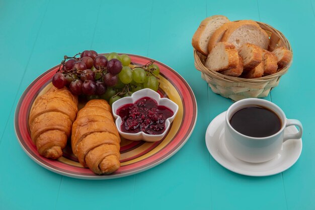 Vista laterale del set colazione con croissant uva marmellata di lamponi e fette di pane con tazza di tè su sfondo blu