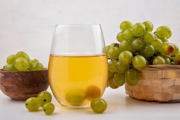 Vista laterale del succo d'uva bianca in vetro con uva bianca nella ciotola e nel cesto su sfondo bianco