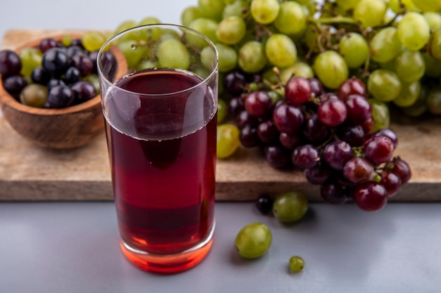 Vista laterale del succo d'uva in vetro e uva con ciotola di acini d'uva sul tagliere su sfondo grigio