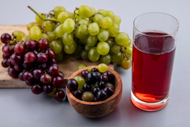 Vista laterale del succo d'uva in vetro e uva sul tagliere con ciotola di acini d'uva su sfondo grigio