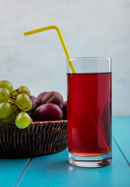 Vista laterale del succo d'uva nera con tubo per bere in vetro e cesto di uva pluots su superficie blu e sfondo bianco
