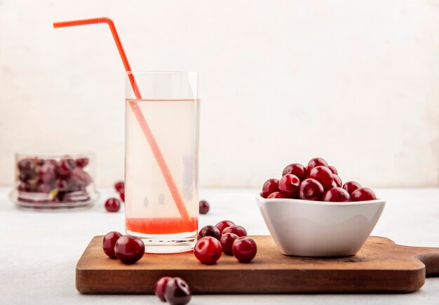 Vista laterale del succo di ciliegia con tubo per bere in vetro e ciliegie nella ciotola e sul tagliere e su sfondo bianco