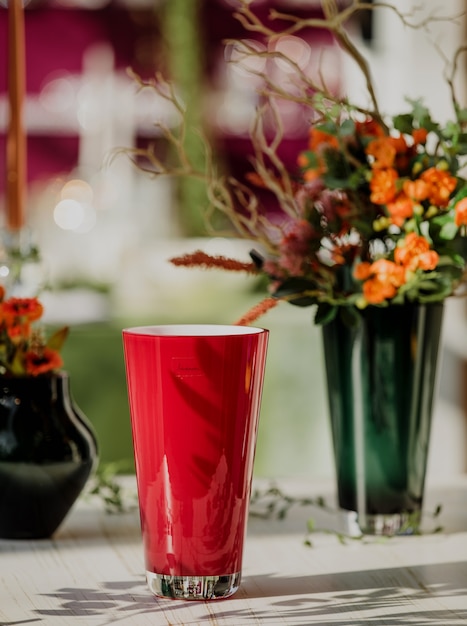 Vista laterale del vetro di colore rosso per acqua o succo sul tavolo con fiori in un vaso sul muro