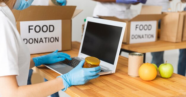 Vista laterale del volontario femminile che tiene cibo di donazione con il computer portatile