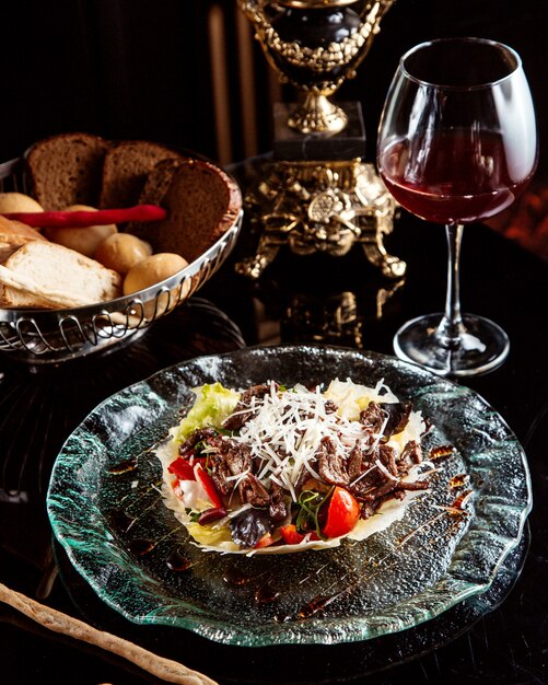 Vista laterale dell'insalata di manzo con verdure e parmigiano su un piatto con vino rosso sul tavolo