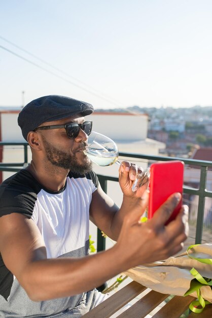 Vista laterale dell'uomo africano felice che tiene smart phone. Uomo romantico in abiti casual e occhiali da sole seduto al tavolo a bere vino guardando il telefono parlando di data. Relazioni umane e concetto di datazione