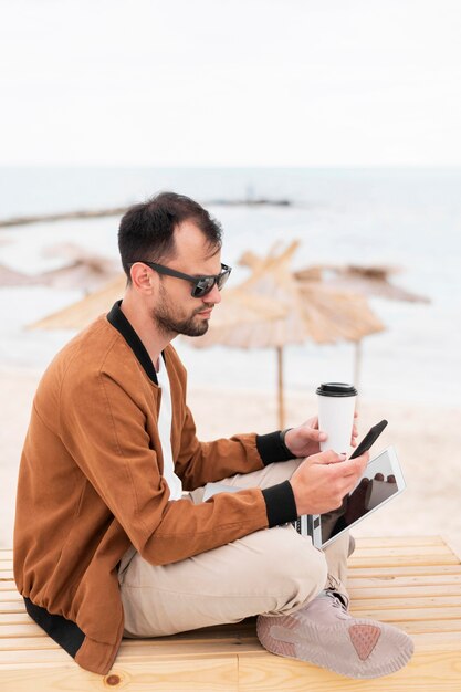 Vista laterale dell'uomo che lavora in spiaggia mentre si mangia il caffè