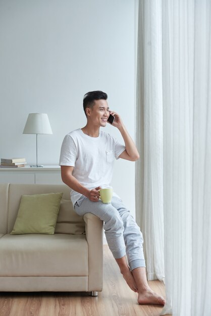 Vista laterale dell'uomo che si appollaia sul bracciolo del divano parlando al telefono e guardando nella finestra