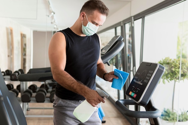 Vista laterale dell'uomo con mascherina medica che disinfetta attrezzature da palestra