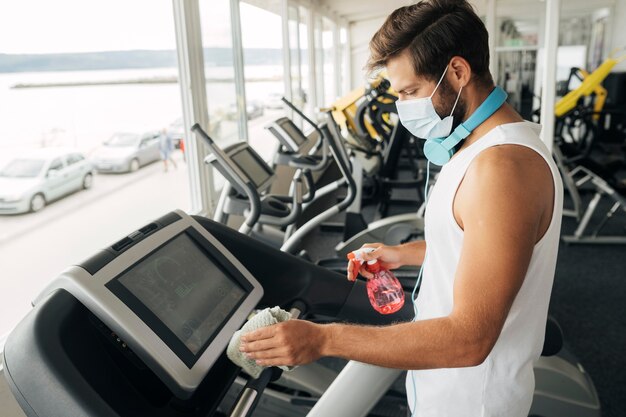 Vista laterale dell'uomo con mascherina medica che disinfetta il tapis roulant in palestra