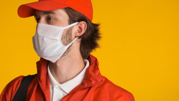 Vista laterale dell'uomo di consegna vestito con uniforme rossa che indossa una maschera protettiva su sfondo colorato Concetto di sicurezza prima di tutto