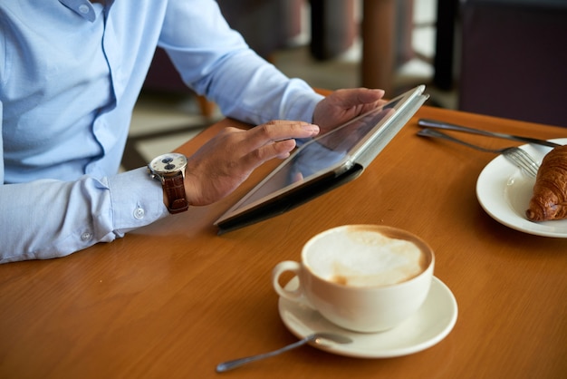 Vista laterale dell'uomo potato che utilizza applicazione mobile sul PC della compressa che mangia caffè con il croissant