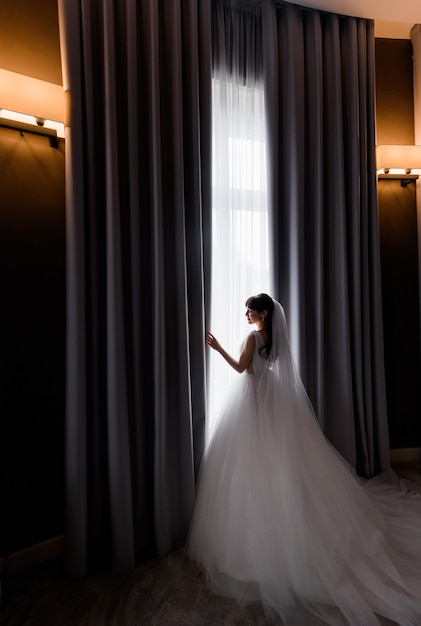 Vista laterale della bella sposa bruna in piedi vicino alla finestra al mattino in una stanza d'albergo buia