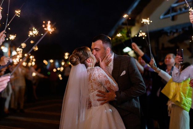 Vista laterale della bella sposa e dello sposo che si abbracciano e si baciano mentre gli ospiti tengono in mano scintillii freddi che bruciano e creano arco luminoso durante la cerimonia notturna all'aperto