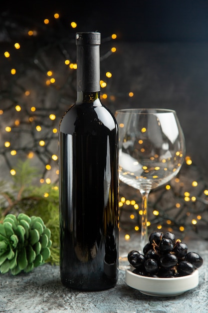 Vista laterale della bottiglia di vino rosso per celebrare un bicchiere vuoto e un cono di conifere di uva nera su sfondo scuro