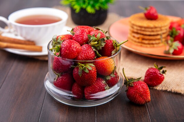 Vista laterale della ciotola di fragola con una tazza di tè e biscotti alla cannella e waffle su tela di sacco e superficie di legno