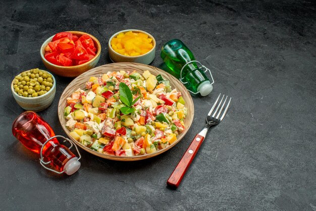 Vista laterale della ciotola di insalata di verdure con una ciotola di forchetta di peperoni tagliati e bottiglia di olio su sfondo scuro