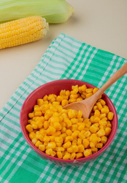 Vista laterale della ciotola di semi del cereale con il cucchiaio sulle pannocchie e pannocchie di granturco sulla tavola bianca