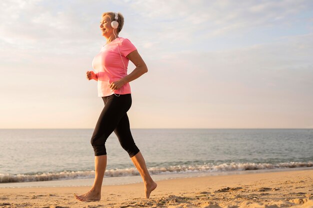 Vista laterale della donna anziana con le cuffie a fare jogging sulla spiaggia