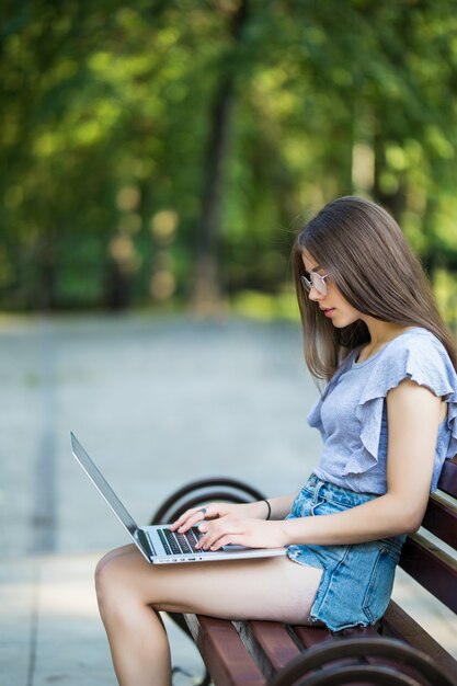 Vista laterale della donna bruna soddisfatta in occhiali che si siede sulla panchina nel parco e utilizzando il computer portatile