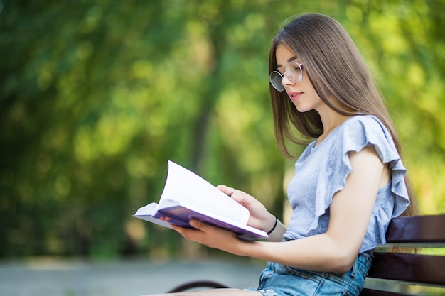 Vista laterale della donna castana lieta in occhiali che si siede sulla panchina e leggere il libro nel parco