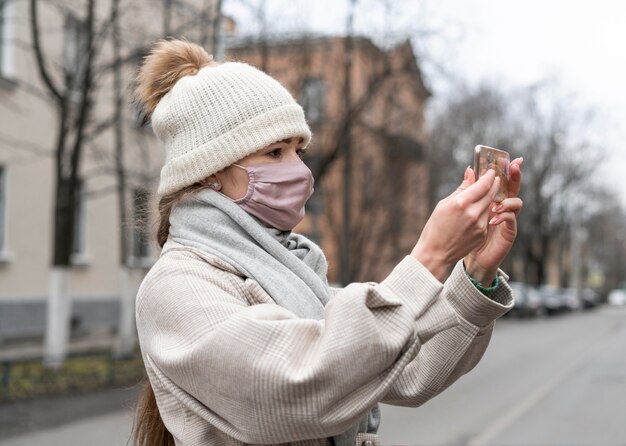 Vista laterale della donna con mascherina medica avente una videochiamata all'aperto