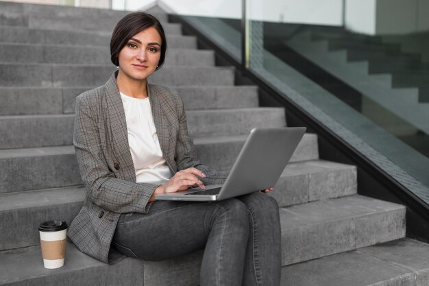 Vista laterale della donna di affari che mangia caffè e lavora al computer portatile sui gradini