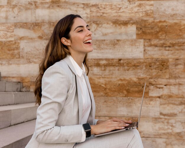 Vista laterale della donna di affari di smiley con smartwatch che lavora al computer portatile all'aperto