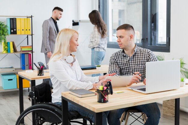 Vista laterale della donna in sedia a rotelle che discute con il collega al suo scrittorio