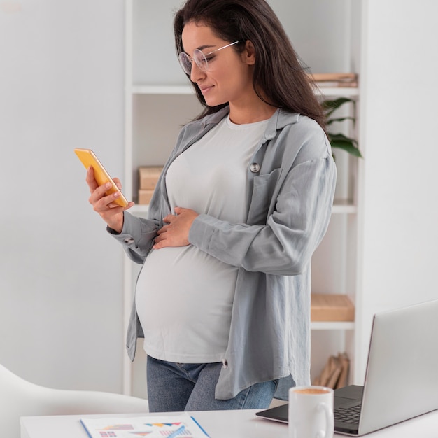 Vista laterale della donna incinta a casa con laptop e smartphone