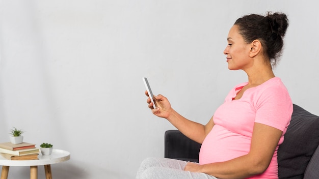 Vista laterale della donna incinta a casa con lo smartphone