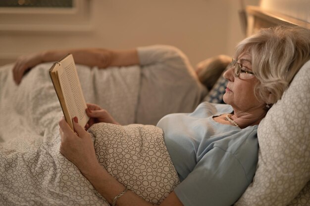 Vista laterale della donna matura sdraiata a letto e leggendo un libro Suo marito sta dormendo sullo sfondo