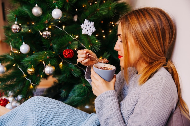 Vista laterale della donna moderna bevendo tè accanto all&#39;albero di Natale