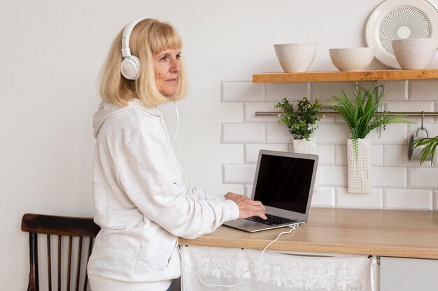 Vista laterale della donna più anziana che ascolta la musica sulle cuffie a casa con il computer portatile