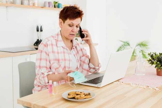 Vista laterale della donna più anziana che parla sulla maschera medica dell'intera tenuta del telefono