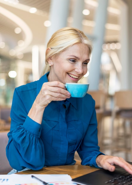 Vista laterale della donna più anziana di affari di smiley che mangia tazza di caffè e che lavora al computer portatile