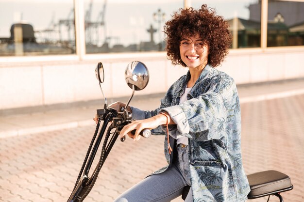 Vista laterale della donna riccia piacevole che si siede sulla motocicletta moderna