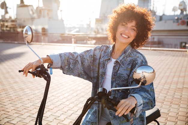 Vista laterale della donna riccia sorridente che si siede sulla motocicletta moderna all'aperto