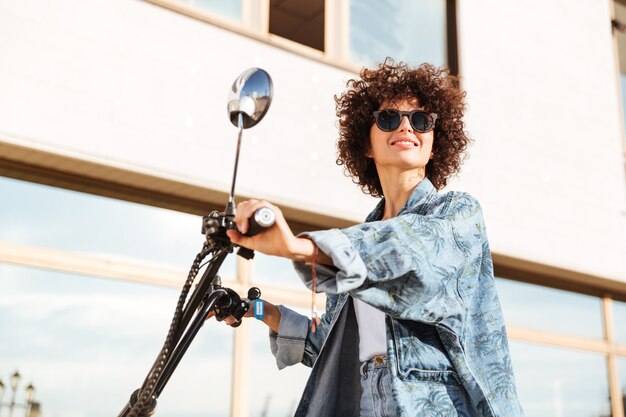 Vista laterale della donna riccia sorridente di bellezza in occhiali da sole che si siedono sul motobike moderno all'aperto e distogliere lo sguardo