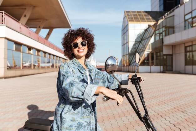 Vista laterale della donna riccia sorridente in occhiali da sole che posano sulla motocicletta moderna all'aperto