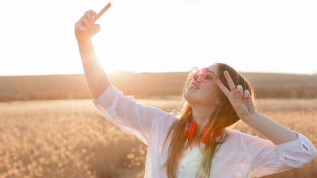 Vista laterale della donna spensierata con gli occhiali da sole che prendono un selfie in natura