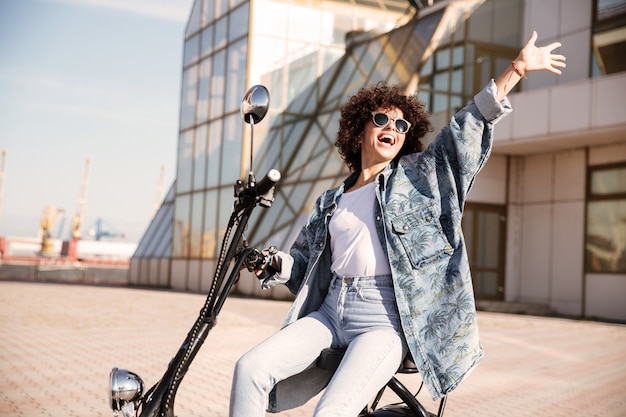 Vista laterale della donna spensierata in occhiali da sole che si siedono sulla moto