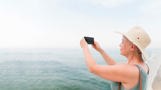 Vista laterale della donna turistica più anziana che prende le immagini sul mare