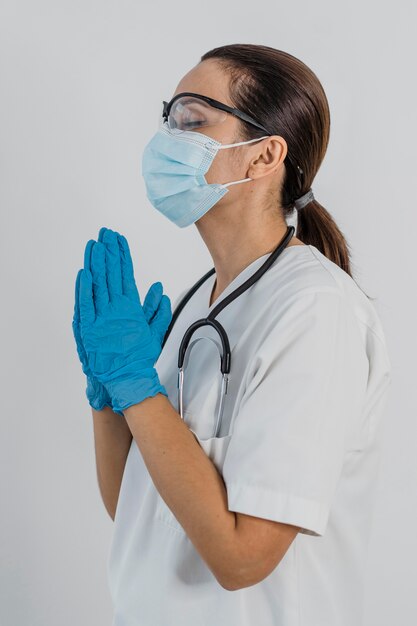 Vista laterale della dottoressa con maschera medica e occhiali di sicurezza pregando