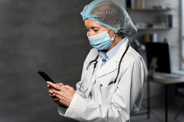 Vista laterale della dottoressa con maschera medica e retina per capelli che tiene smartphone