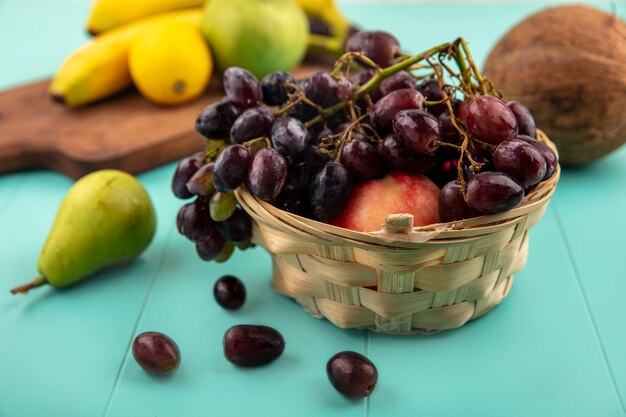 Vista laterale della frutta come cesto di uva e pesca con banana mela limone sul tagliere e pera cocco su sfondo blu