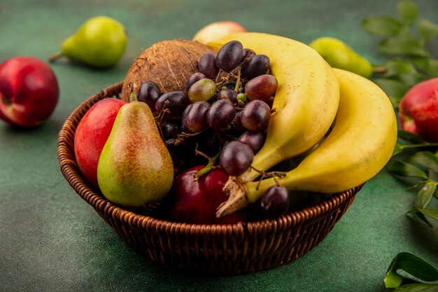 Vista laterale della frutta come merce nel cestino della pesca della pera dell'uva della banana della noce di cocco con i fogli su fondo verde