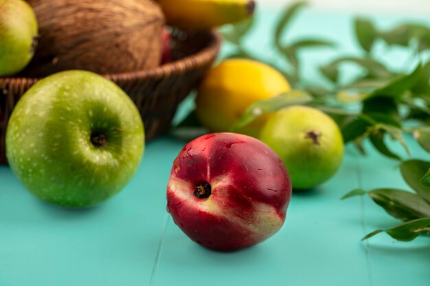 Vista laterale della frutta come pesca mela pera limone con cesto di cocco banana e foglie su sfondo blu